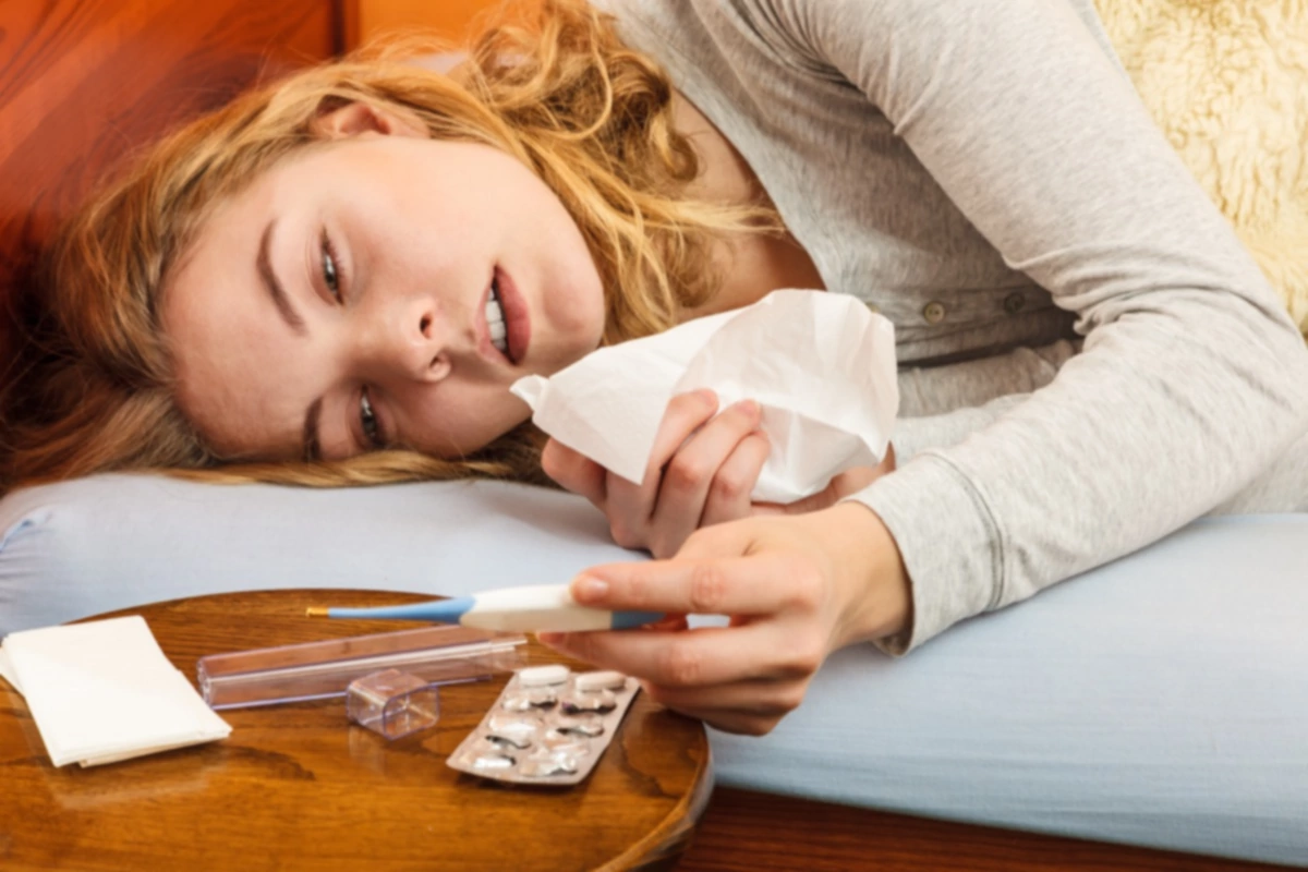 Mujer enferma tumbada en la cama bajo una manta de lana sosteniendo un termómetro y un pañuelo. Niña enferma que ha cogido un resfriado. Píldoras y pastillas sobre la mesa.