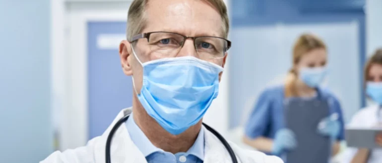 Viejo médico maduro con bata blanca, estetoscopio, gafas y mascarilla de pie en un hospital mirando a la cámara.